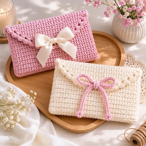 May include: Two handmade crochet envelope-style pouches. One is light pink with a cream-colored bow, the other is off-white with a pink bow. Both have scalloped edges and are displayed on a wooden tray with flowers.