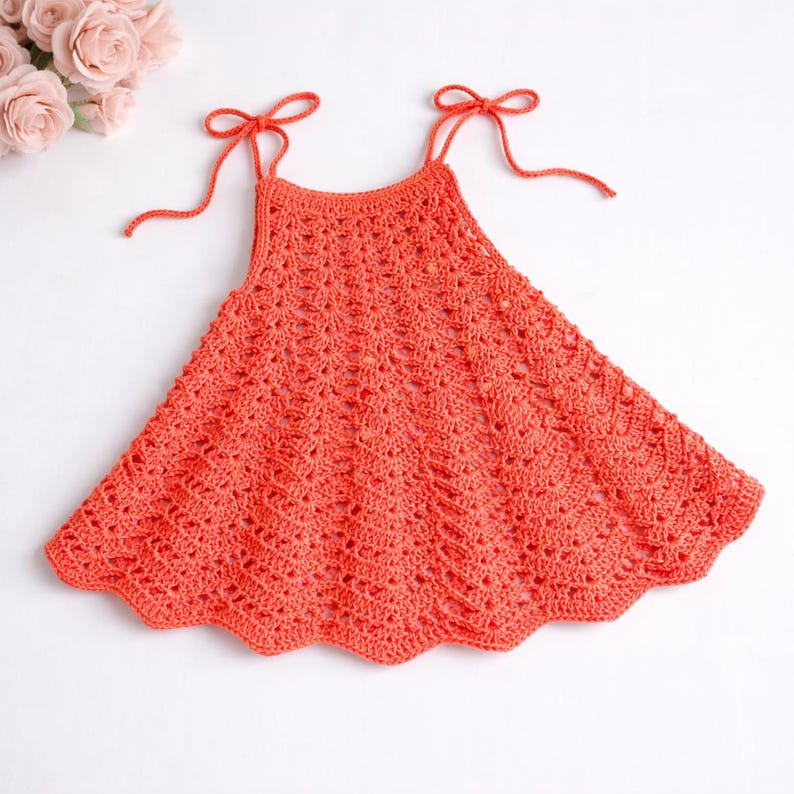May include: Coral-colored crochet dress with tie straps. The dress features a scalloped hemline and a textured, openwork design. The straps are tied into bows. The dress is likely handmade and suitable for a child.
