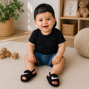 May include: A black baseball cap, a black short-sleeved t-shirt with a chest pocket, blue denim shorts, and black crocheted sandals featuring a white swoosh-like emblem on the top strap. The outfit is worn by a young child with a smiling expression.