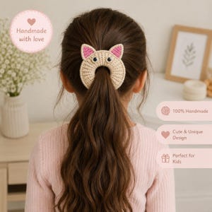 Könnte beinhalten: Ein beiges Häkel-Haaraccessoire in Form eines Katzenkopfes mit rosa Innenohren und zwei schwarzen Kunststoffaugen. Das kreisförmige Design verfügt über eine zentrale Öffnung zum Befestigen der Haare und ist mit detaillierten Maschen gefertigt.