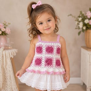 May include: A handmade crochet top with a white and pink floral design. The top has a square neckline and straps, paired with a white eyelet skirt. A pink bow is in the child's hair. The outfit is perfect for a summer day.