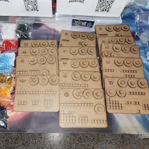 Op de afbeelding: Een verzameling lasergesneden bruine houten spelborden met ronde sleuven en gegraveerde volggrids, vergezeld door verschillende kleine plastic zakjes gevuld met diverse gekleurde doorschijnende acrylblokjes in rood, geel, blauw, groen en zwart.