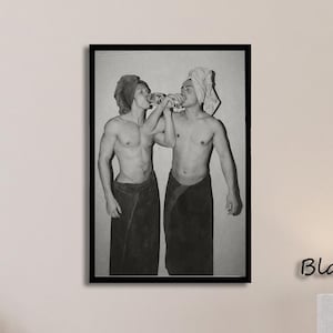 May include: A black and white framed wall art print featuring two individuals wearing towels wrapped around their waists and heads, standing side by side while drinking from a shared bottle. The artwork is enclosed in a simple black frame.
