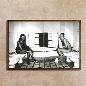 Op de afbeelding: Een zwart-witfoto in een notenhouten lijst met twee personen die op toiletten zitten in een betegelde badkamer. De afbeelding is in het midden gespleten en toont &eacute;&eacute;n persoon aan de linkerkant en een andere aan de rechterkant, met witte handdoeken.