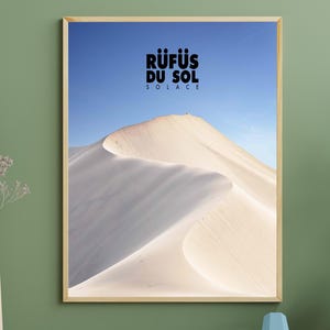 May include: A framed print featuring a desert landscape with a sand dune under a clear blue sky. The print has the text "RÜFÜS DU SOL SOLACE" in black at the top. The frame is a light wood color.