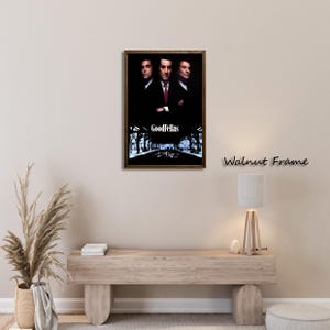 May include: A framed movie poster featuring three men in suits, with the title "Goodfellas" at the bottom. The frame is a dark walnut color. The poster is displayed on a light-colored wall above a wooden bench.