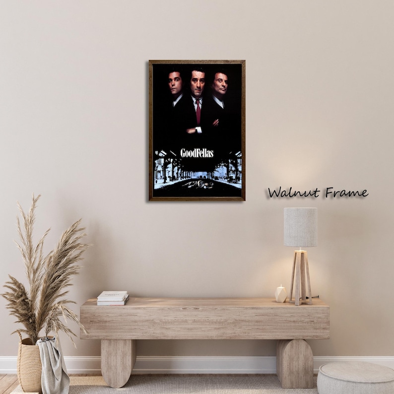 May include: A framed movie poster featuring three men in suits, with the title "Goodfellas" at the bottom. The frame is a dark walnut color. The poster is displayed on a light-colored wall above a wooden bench.