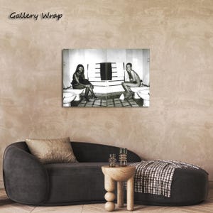 Op de afbeelding: Een zwart-wit gallery wrap canvas print met twee personen zittend op toiletten in een betegelde badkamer. Het kunstwerk toont een gespleten compositie met een centraal handdoekenrek en witte sanitaire voorzieningen in een monochroom contrast.