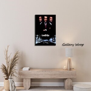 May include: A framed canvas print of the movie "Goodfellas." The artwork features three men in suits against a dark background, with the film's title at the bottom. The print is mounted on a wall above a wooden console.