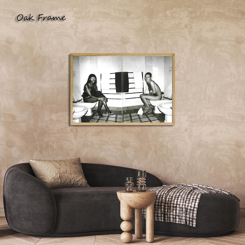 May include: Black and white framed print featuring two figures seated on toilets. The artwork is displayed in a light oak frame and hangs above a dark gray sofa and a wooden side table. The text "Oak Frame" is visible in the upper left corner.