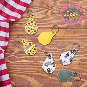 May include: A collection of keychains in various designs, including sunflowers, a yellow solid color, and a black and white pattern. The keychains are on a wooden surface with a red and white checkered cloth in the corner. The text "HOBBY JOBBY CREATIONS" is visible.