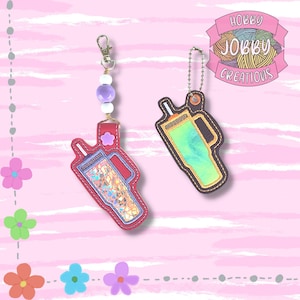 Könnte beinhalten: Zwei Getränke-förmige Schlüsselanhänger. Einer ist rot mit einem Blumenakzent und schillerndem Glitzer. Der andere ist braun mit einem grün-gelben Design. Beide haben eine genähte Kontur und einen Metallverschluss oder eine Kette.