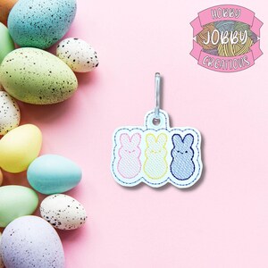 May include: A white keychain with three embroidered bunny figures in pink, yellow, and blue. The keychain is attached to a metal clasp. The background is pink with colorful Easter eggs.