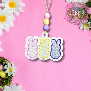May include: An Easter-themed hanging ornament featuring three embroidered bunny figures in pink, yellow, and blue. The ornament is attached to a string of decorative beads in coordinating colors. The background is a soft pink, with floral accents.