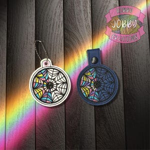 May include: Two round keychains with a stained glass spiderweb design. One is white with a silver chain, the other is navy blue with a snap closure. The spiderwebs are multicolored with black outlines. The logo "Hobby Jobby Creations" is visible.
