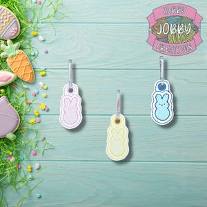May include: Three Easter bunny-shaped zipper pulls in pink, yellow, and blue, each with a silver clasp. The background is a light blue wooden surface with Easter-themed cookies and decorative grass. The logo "HOBBY JOBBY CREATIONS" is in the upper right.
