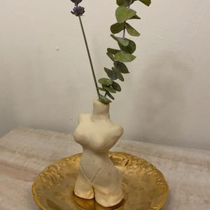 May include: A cream-colored ceramic vase in the shape of a torso, holding sprigs of lavender and eucalyptus. The vase sits on a gold-colored, ornate plate. The arrangement is set against a neutral background.