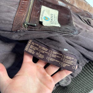 K&ouml;nnte beinhalten: Nahaufnahme einer braunen Lederjacke mit Rei&szlig;verschluss und einem Etikett mit der Aufschrift "REDKINS EQUIPMENT". Die Jacke hat ein dunkelbraunes Futter und ein geripptes B&uuml;ndchen. Das Etikett tr&auml;gt auch den Text "LIGHTSQUADRON".