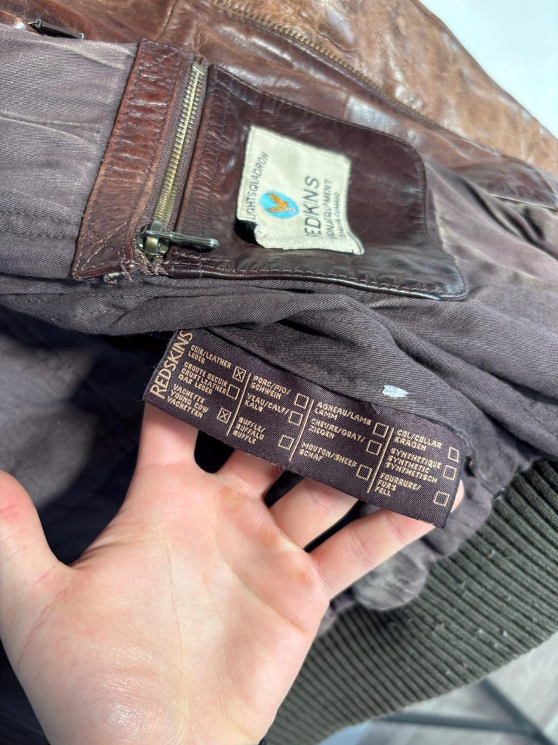 K&ouml;nnte beinhalten: Nahaufnahme einer braunen Lederjacke mit Rei&szlig;verschluss und einem Etikett mit der Aufschrift "REDKINS EQUIPMENT". Die Jacke hat ein dunkelbraunes Futter und ein geripptes B&uuml;ndchen. Das Etikett tr&auml;gt auch den Text "LIGHTSQUADRON".