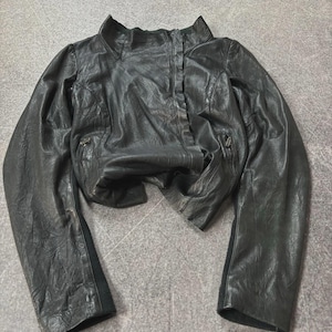 May include: A black leather jacket with a stand-up collar and long sleeves. The jacket has a cropped design and features two zippered pockets. The leather appears worn and textured, giving it a vintage look.