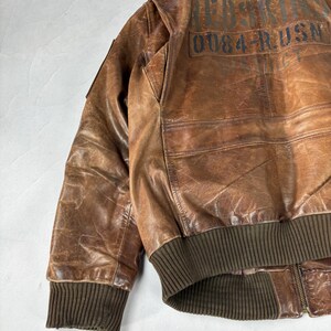 K&ouml;nnte beinhalten: Braune Leder-Bomberjacke mit gerippten B&uuml;ndchen und Bund. Auf der R&uuml;ckseite der Jacke befindet sich aufgedruckter Text, darunter "RED SKIN" und "0084-RUSN". Die Jacke hat einen Rei&szlig;verschluss und einen klassischen Bomberstil.