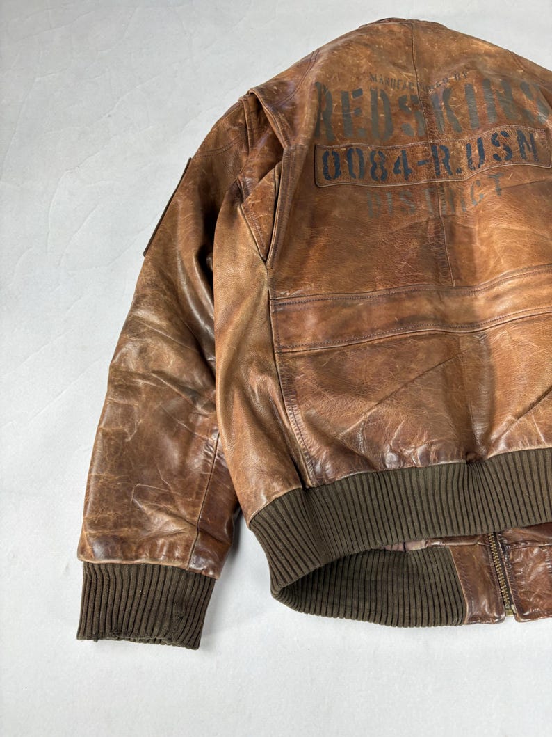 K&ouml;nnte beinhalten: Braune Leder-Bomberjacke mit gerippten B&uuml;ndchen und Bund. Auf der R&uuml;ckseite der Jacke befindet sich aufgedruckter Text, darunter "RED SKIN" und "0084-RUSN". Die Jacke hat einen Rei&szlig;verschluss und einen klassischen Bomberstil.
