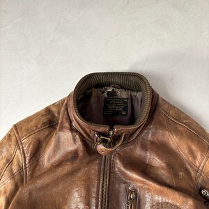 K&ouml;nnte beinhalten: Braune Lederjacke mit Rei&szlig;verschluss vorne, geripptem Kragen und kleiner Rei&szlig;verschlusstasche auf der Brust. Die Jacke hat ein abgenutztes Aussehen und einen Logoaufn&auml;her auf der Brust. Die Jacke ist im Vintage-Stil.