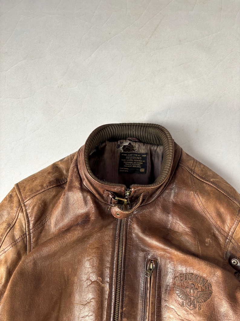 K&ouml;nnte beinhalten: Braune Lederjacke mit Rei&szlig;verschluss vorne, geripptem Kragen und kleiner Rei&szlig;verschlusstasche auf der Brust. Die Jacke hat ein abgenutztes Aussehen und einen Logoaufn&auml;her auf der Brust. Die Jacke ist im Vintage-Stil.