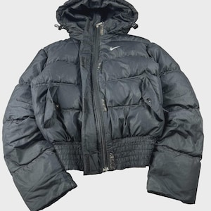Op de afbeelding: Een zwart pufferjack met een capuchon met verstelbare trekkoorden, een ritssluiting over de hele lengte en een geribbelde elastische tailleband. Het jack heeft twee zijzakken met drukknopen en een klein wit Nike-logo op de borst.