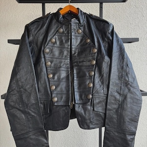 May include: Black leather jacket with a military-style design. The jacket features multiple decorative buttons, a zipper closure, and a mandarin collar. The jacket is hanging on a wooden hanger.