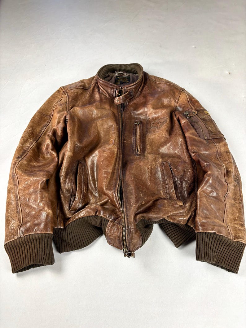 K&ouml;nnte beinhalten: Braune Leder-Bomberjacke mit geripptem Kragen, B&uuml;ndchen und Saum. Die Jacke hat einen Rei&szlig;verschluss, mehrere Taschen und ein Used-Finish. Die Ma&szlig;e der Jacke sind auf dem Bild nicht sichtbar.