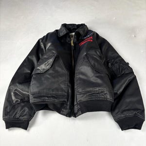 Peut inclure: Veste de bombardier noire avec une fermeture éclair, deux poches avant et un ourlet et des poignets côtelés. Le mot "ABSOLUTEX" est écrit en rouge sur la poitrine. La veste est faite d'un matériau brillant semblable à du satin.