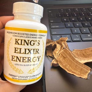 May include: A white supplement bottle with a yellow label featuring the text "KING'S ELIXIR ENERGY" and the words "Mushroom Boosted Energy Formula." The bottle is held near a laptop and a piece of dried mushroom.