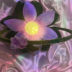 Puede incluir: Una diadema floral con una flor grande de color púrpura claro con un centro amarillo brillante. La flor está rodeada de hojas verdes y una diadema verde. Un pequeño grupo de flores moradas adorna la diadema.