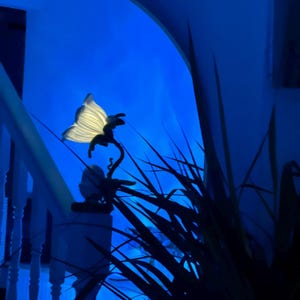 Op de afbeelding: Een decoratieve vlindervormige lamp met een wit, oplichtend lichaam en donkere silhouetdetails. De lamp is gemonteerd op een gebogen steel, tegen een helderblauwe achtergrond. De afbeelding bevat een witte trap en donkergroen gebladerte.