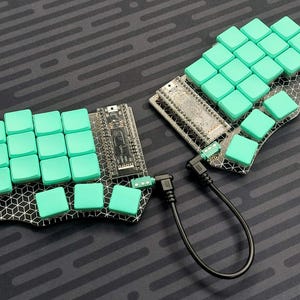 Op de afbeelding: Gesplitst ergonomisch toetsenbord met mintgroene keycaps. Het toetsenbord is in twee helften gesplitst, verbonden door een zwarte kabel. De keycaps zijn vierkant en gerangschikt in een rasterpatroon. De basis heeft een geometrisch ontwerp.