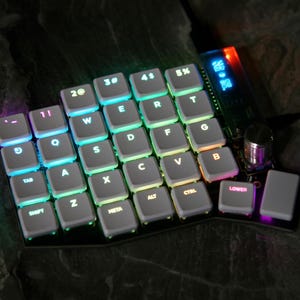 Può includere: Una tastiera ergonomica personalizzata con tasti retroilluminati. I tasti sono disposti in un layout diviso con una varietà di colori. La tastiera ha un piccolo schermo e un encoder rotativo. I tasti sono etichettati con lettere, numeri e simboli.