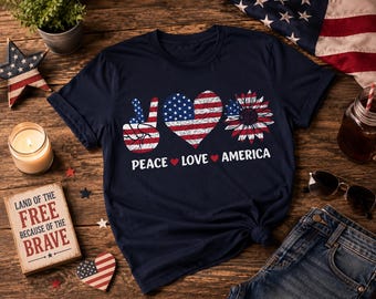 May include: A navy blue short-sleeve t-shirt featuring a graphic print with a peace sign, a heart, and a sunflower, all patterned with the American flag. Below the graphic, the text reads PEACE LOVE AMERICA, separated by small red hearts.