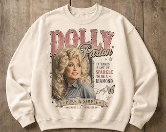 May include: A cream-colored crewneck sweatshirt featuring a graphic print of Dolly Parton. The design includes the text DOLLY Parton, IT TAKES A LOT OF SPARKLE TO BE A DIAMOND, PURE & SIMPLE, and NASHVILLE, TENNESSEE, along with a butterfly illustration.