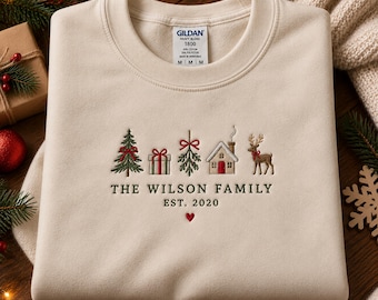 May include: A cream-colored crewneck sweatshirt featuring embroidered Christmas icons including a tree, gift, mistletoe, house, and reindeer. Below the icons, the text reads THE WILSON FAMILY EST. 2020 with a small red heart centered underneath.