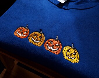 May include: A royal blue crewneck sweatshirt featuring an embroidered design of four jack-o'-lanterns in varying shades of orange and yellow. Each pumpkin displays a unique carved face with stitched details, positioned horizontally across the chest.