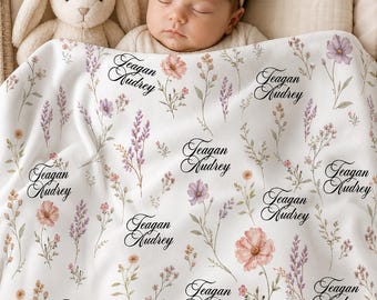 May include: A baby rests under a white blanket with a floral pattern and the name "Teagan Audrey" in black script. The blanket features pink, purple, and yellow flowers. A stuffed bunny and a baby with a bow headband are also in the image.