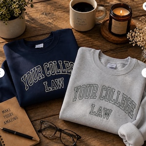 May include: Two sweatshirts, one navy blue and one gray, are displayed on a wooden surface. Both sweatshirts have the text "YOUR COLLEGE LAW" in a collegiate font. A coffee mug, a candle, and a notebook are also visible.