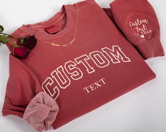 May include: A maroon sweatshirt with the word "CUSTOM" in white block letters. The sleeve cuff has the text "Custom Text Year". A gold necklace with three name pendants and a heart is on the sweatshirt.
