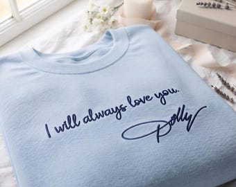 May include: A light blue crewneck sweatshirt featuring dark blue embroidered text that reads I will always love you. followed by the signature Dolly in a cursive script font. The garment has a soft texture and a classic round neckline design.
