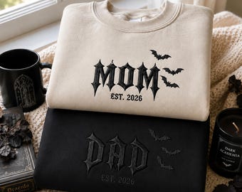 May include: A beige sweatshirt with black embroidered text reading MOM EST. 2026 and three bats, stacked on a black sweatshirt with black embroidered text reading DAD EST. 2026 and three bats. Both garments feature a gothic font style.
