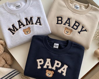 May include: Three crewneck sweatshirts featuring embroidered text and a small bear face graphic. One light gray sweatshirt with dark navy text reading MAMA, one beige sweatshirt with dark navy text reading BABY, and one navy sweatshirt with beige text reading PAPA.