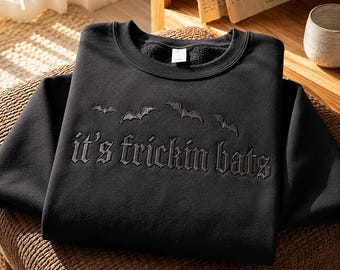 May include: A black crewneck sweatshirt featuring dark grey embroidery of four flying bats arranged in an arc above the text "it's frickin bats" written in a gothic-style font. The garment has long sleeves and a ribbed collar neckline.