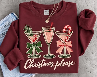 May include: A maroon crewneck sweatshirt featuring a graphic of three cocktail glasses decorated with a rosemary sprig, a candy cane, and bows. The text "Christmas, please" is printed in white script below the glasses. A gold chain necklace rests on the collar.