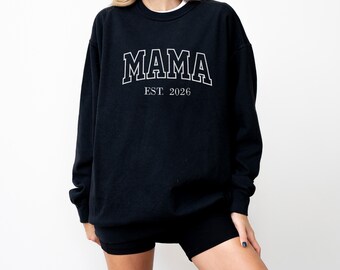 May include: Black sweatshirt with the word "MAMA" in white, outlined block letters. Below the word "MAMA" is the text "EST. 2026" in a smaller font. The sweatshirt is a long-sleeved crew neck.
